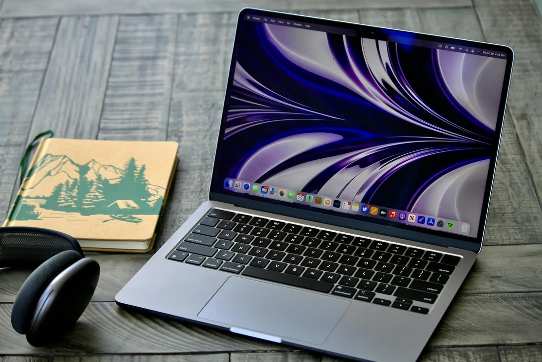 MacBook aberto em uma mesa de madeira, com fones e um caderno ao lado